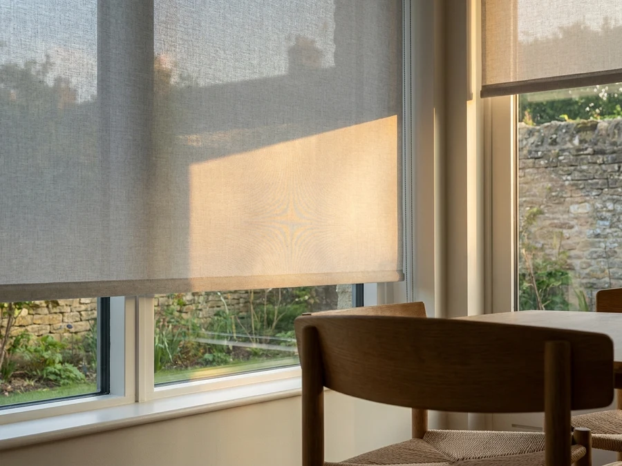 Pale grey roller blind partly lowered across a dining-room window catching warm evening light